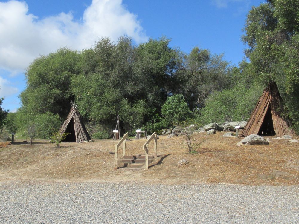 This is a photograph of a typical Chuckchansi and Yokut Native American village. It shows two teepees made of cedar bark, a firepit and other structures for processing acorns.